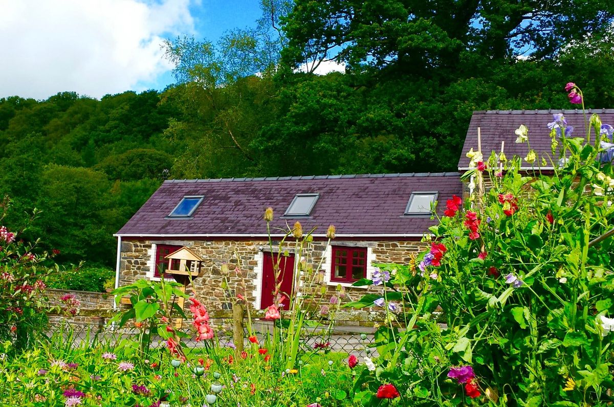 Old Oak Barn, a stone barn with red doors, seen across a garden full of flowers